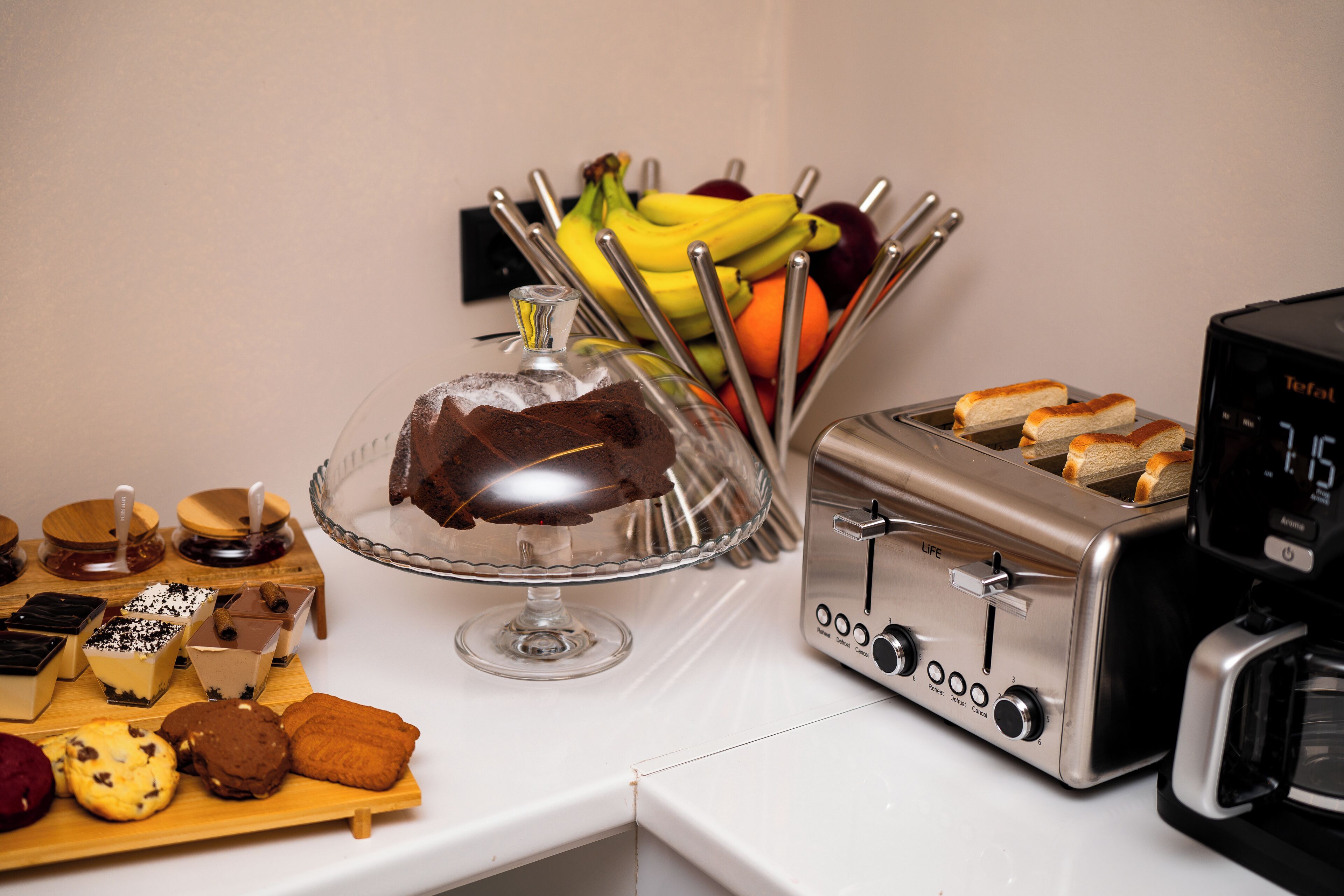 A kitchen counter with a cake on a glass stand, a toaster, and a fruit basket.