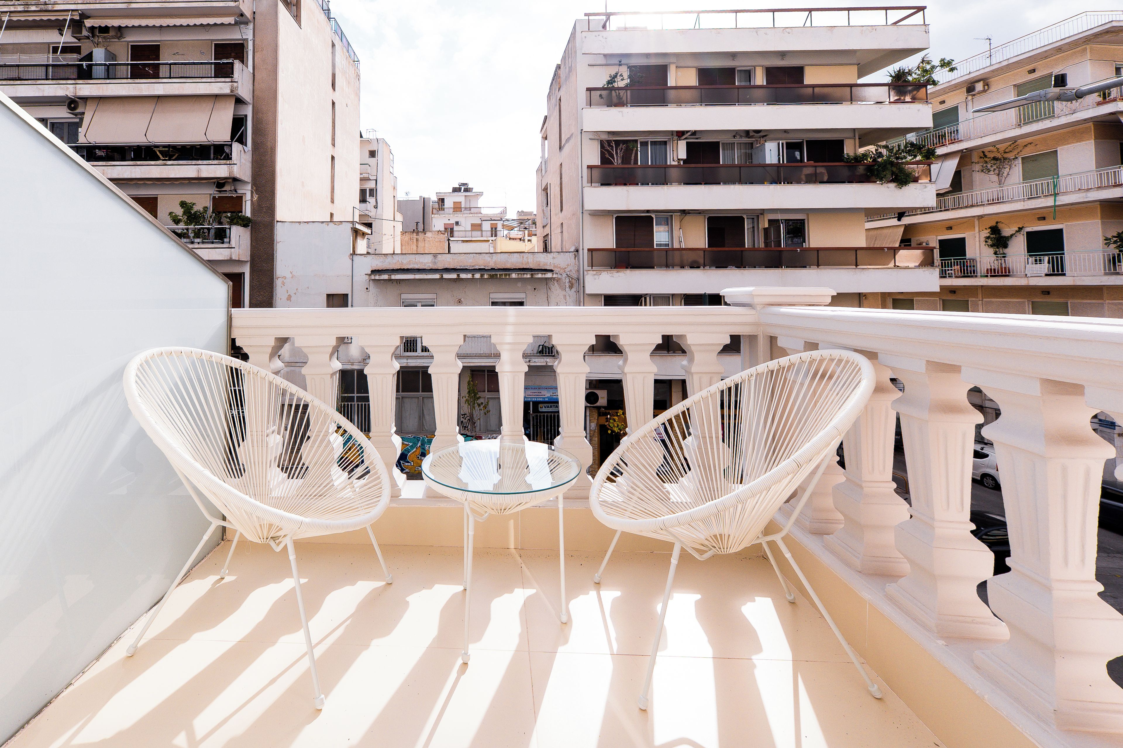 A balcony with two white wicker chairs and a small round table, overlooking a cityscape with multi-story buildings.