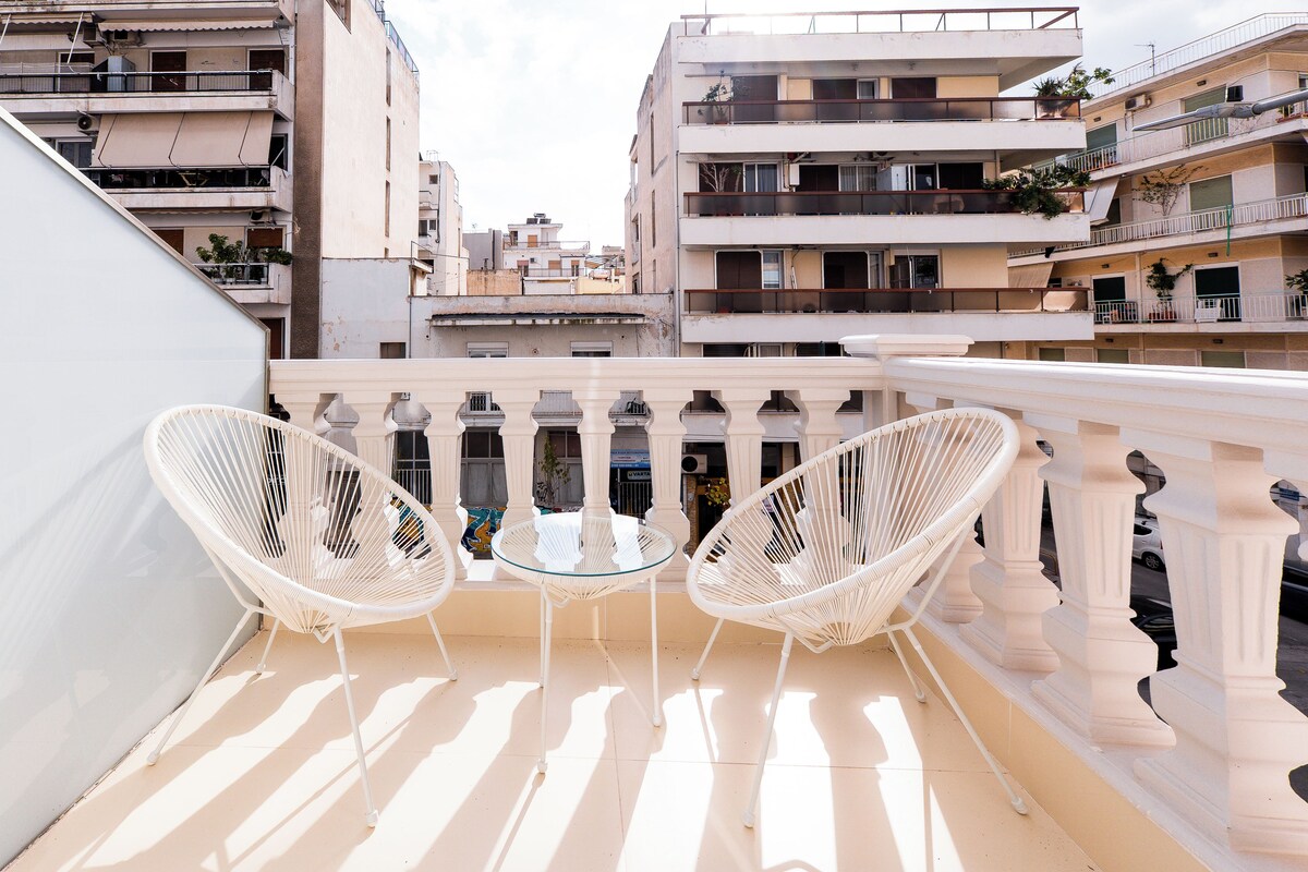 A balcony with two white wicker chairs and a small round table, overlooking a cityscape with multi-story buildings.