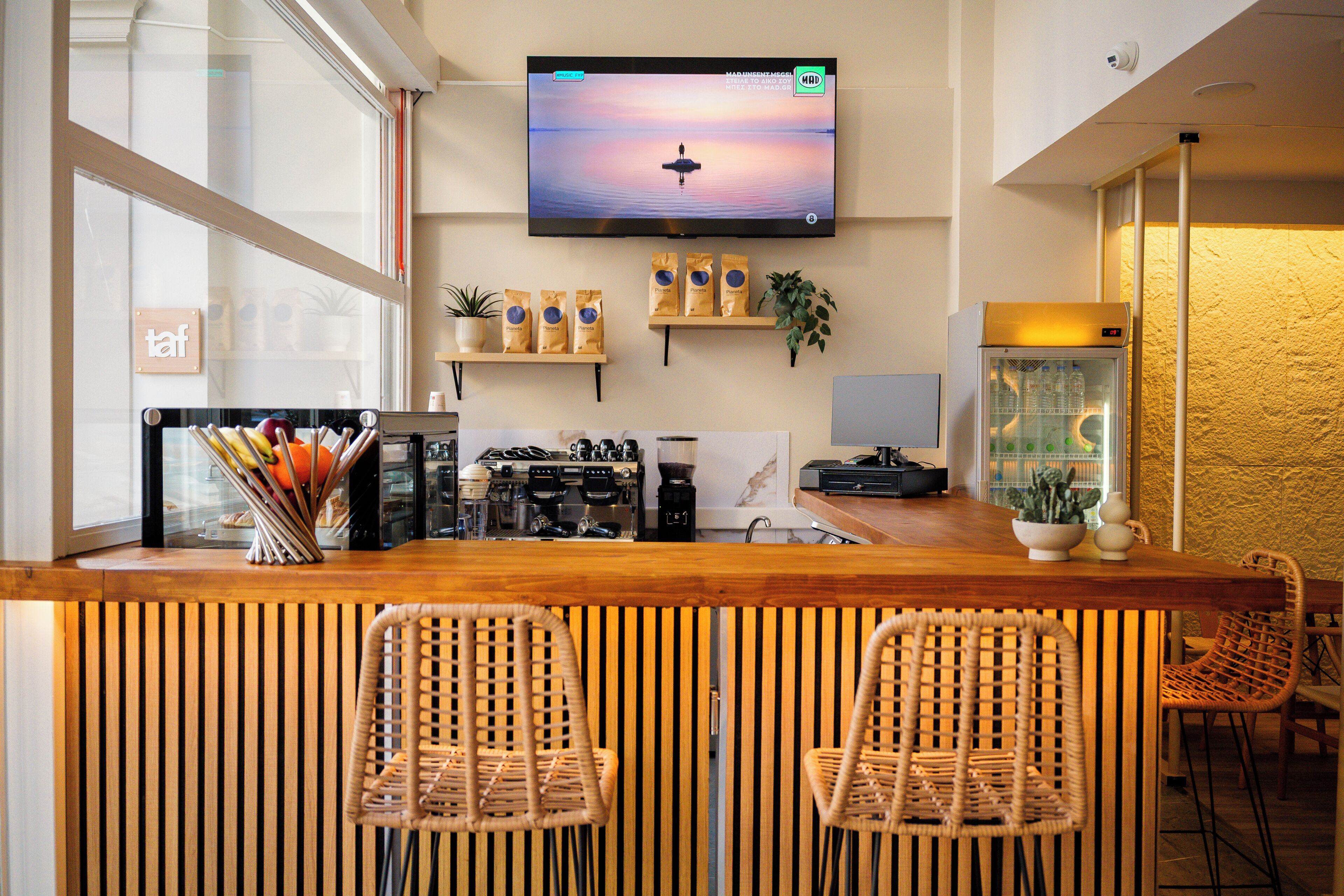 Una zona de bar con mostrador de madera, dos sillas de mimbre, un televisor montado en la pared y un refrigerador con botellas.