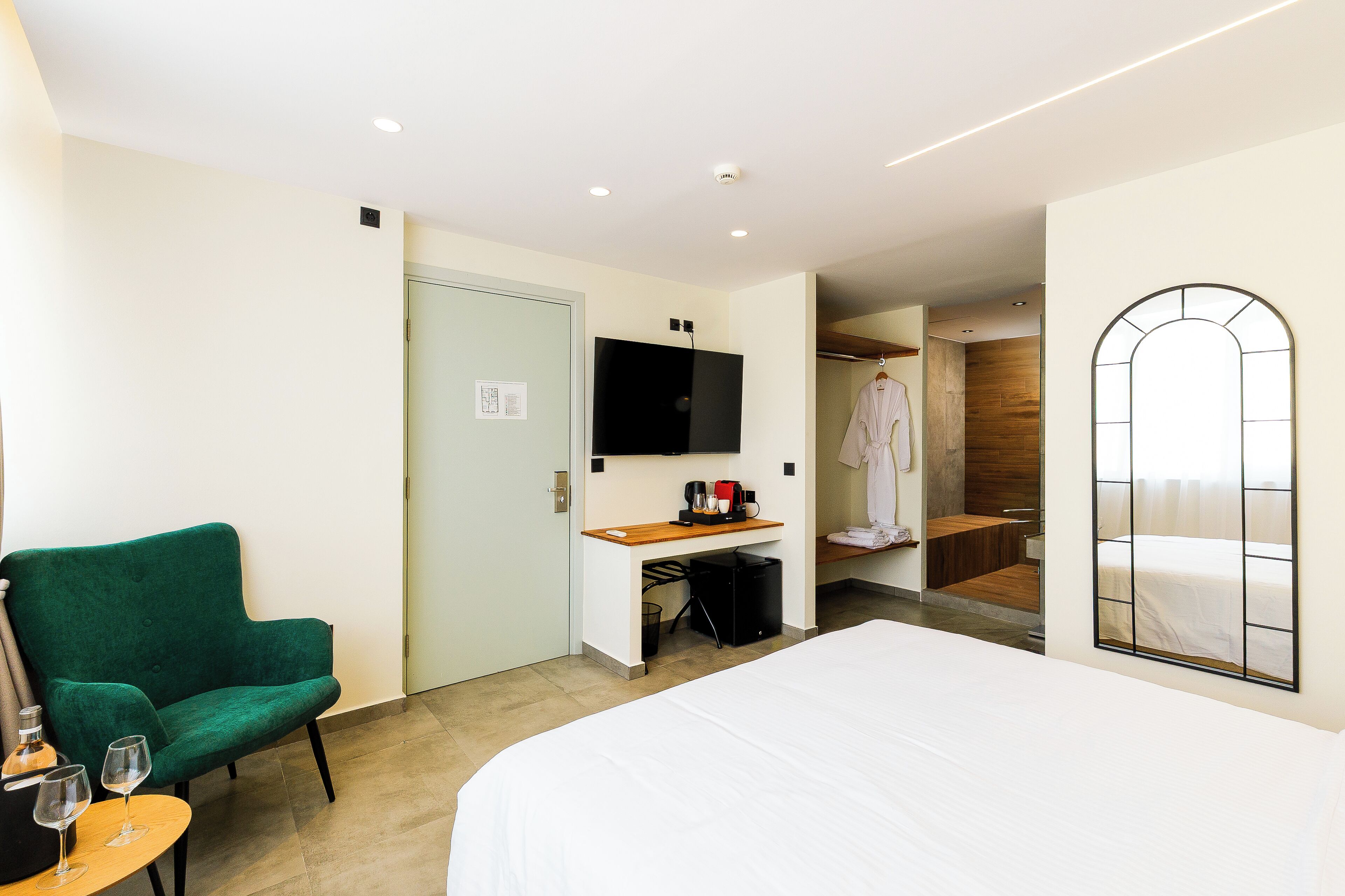 Habitación de hotel con cama, un sillón verde, una mesita con vasos, cafetera, televisor y espejo.