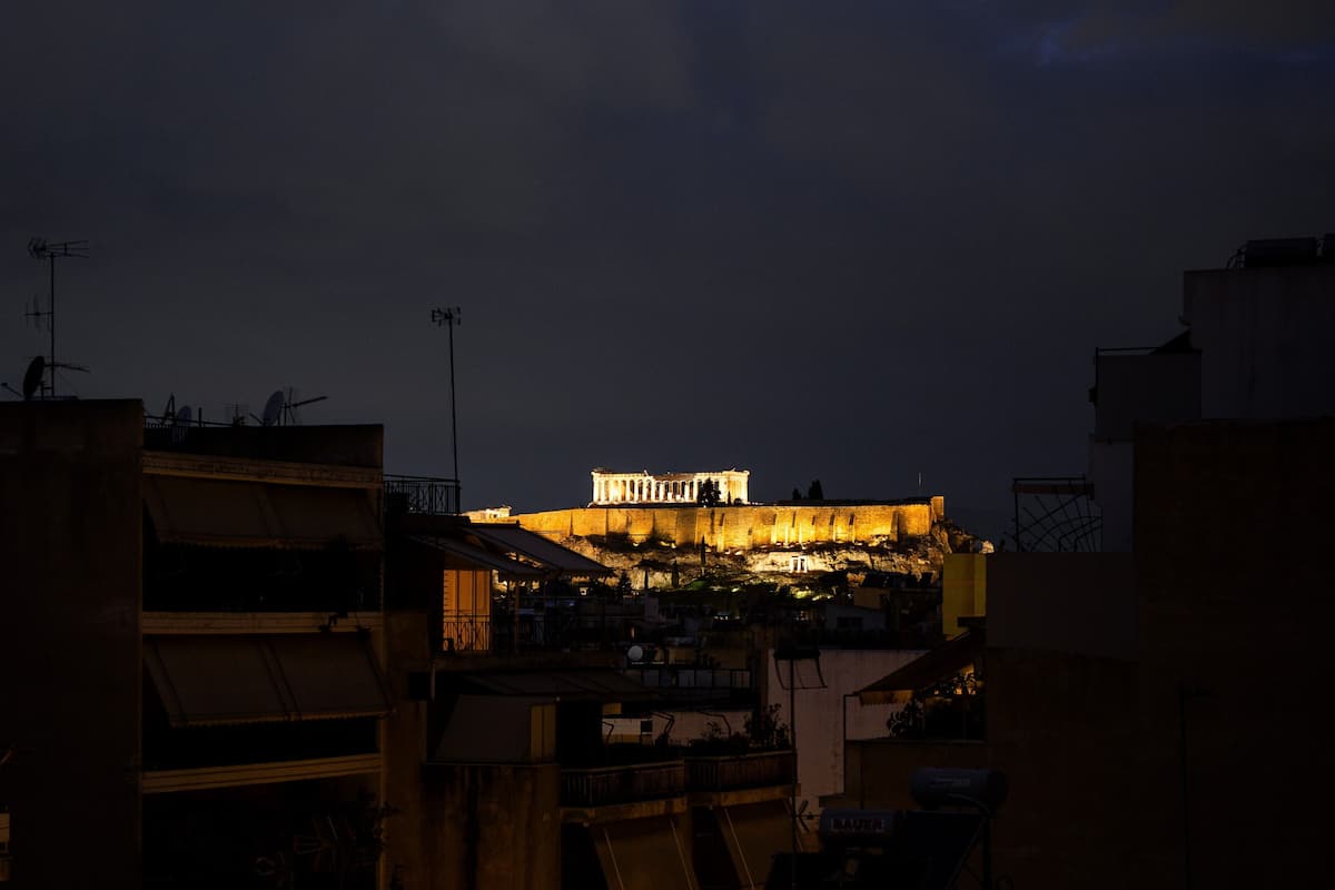 Um edifício histórico com colunas iluminadas e uma entrada imponente, rodeado por edifícios modernos.