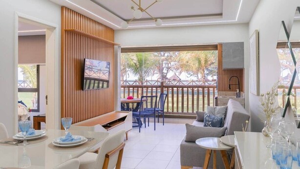 Un soggiorno moderno con angolo pranzo, TV a parete e un balcone con vista su palme.