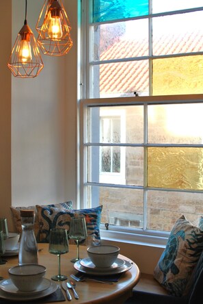 A dining area with a table set for four, a window with a view of a brick building, and hanging pendant lights. - Fisherman's Loft- Seafront Serenity (Anstruther)