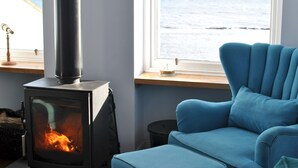 Una habitación con estufa de leña, un sillón azul y ventana con vista al mar.