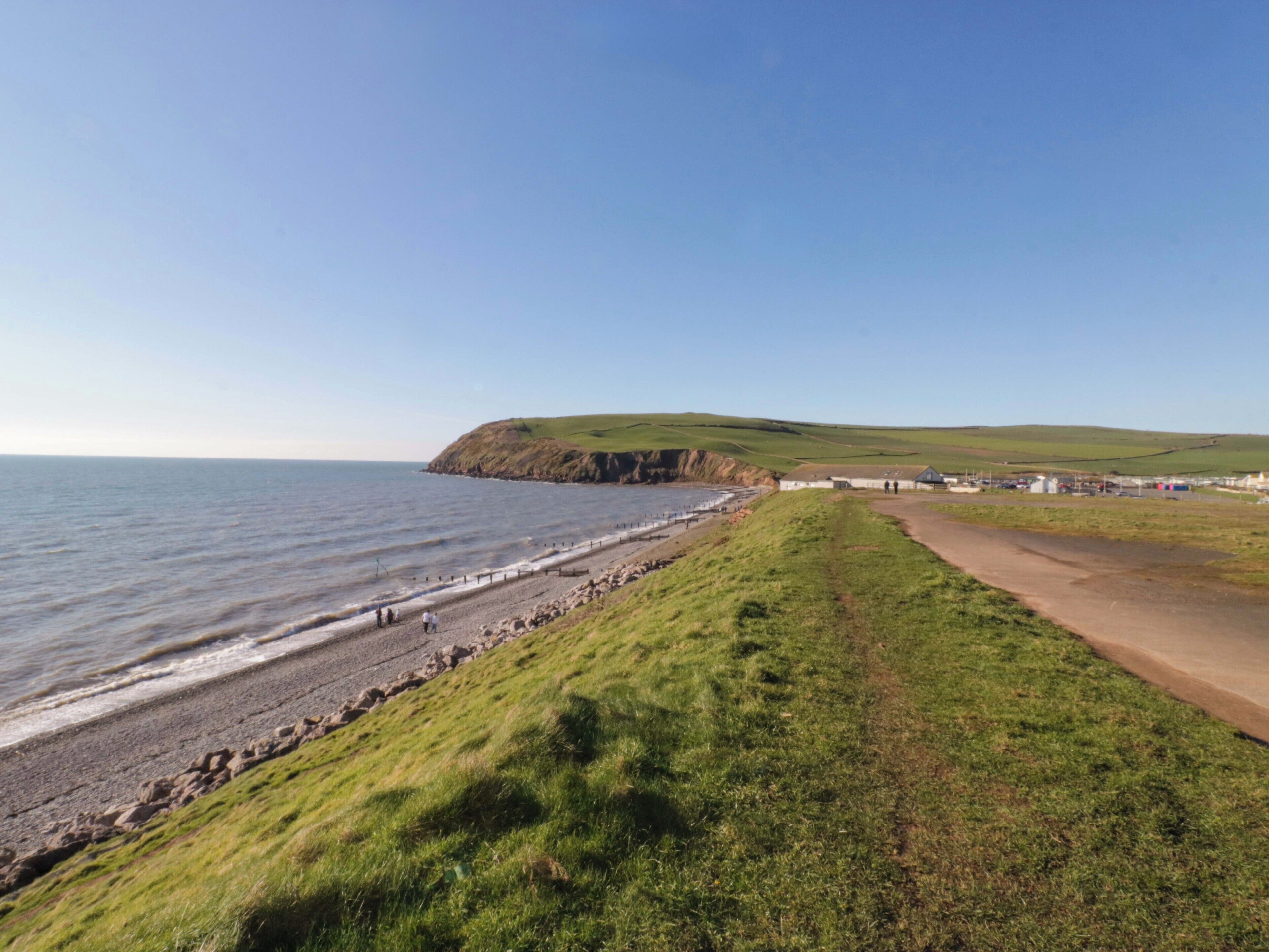 A coastal road leading to a beach with a grassy embankment and a cliff in the background.