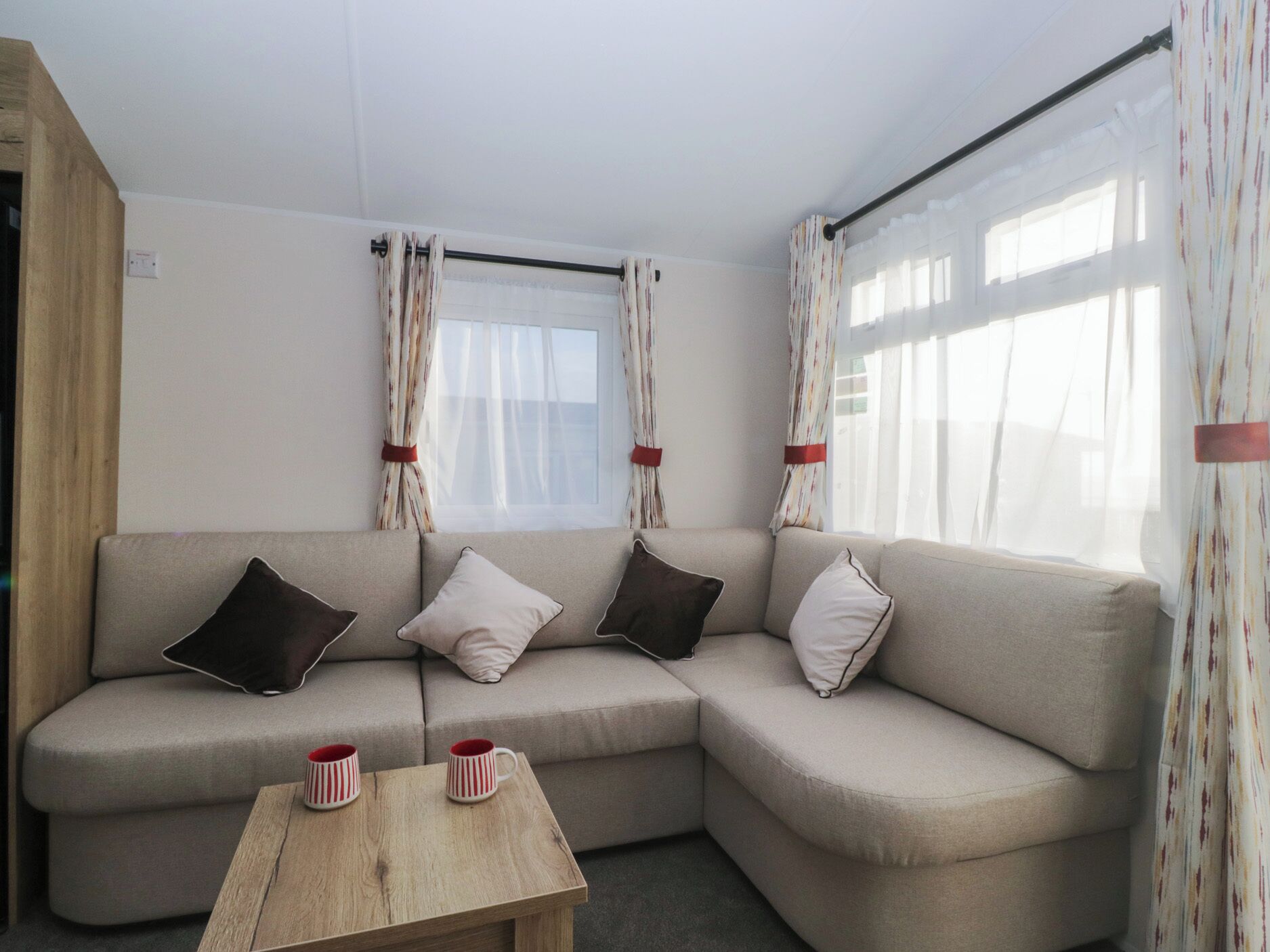 Un sofá de esquina con dos cojines, una mesa de madera con dos tazas a rayas y una ventana con cortinas traslúcidas.