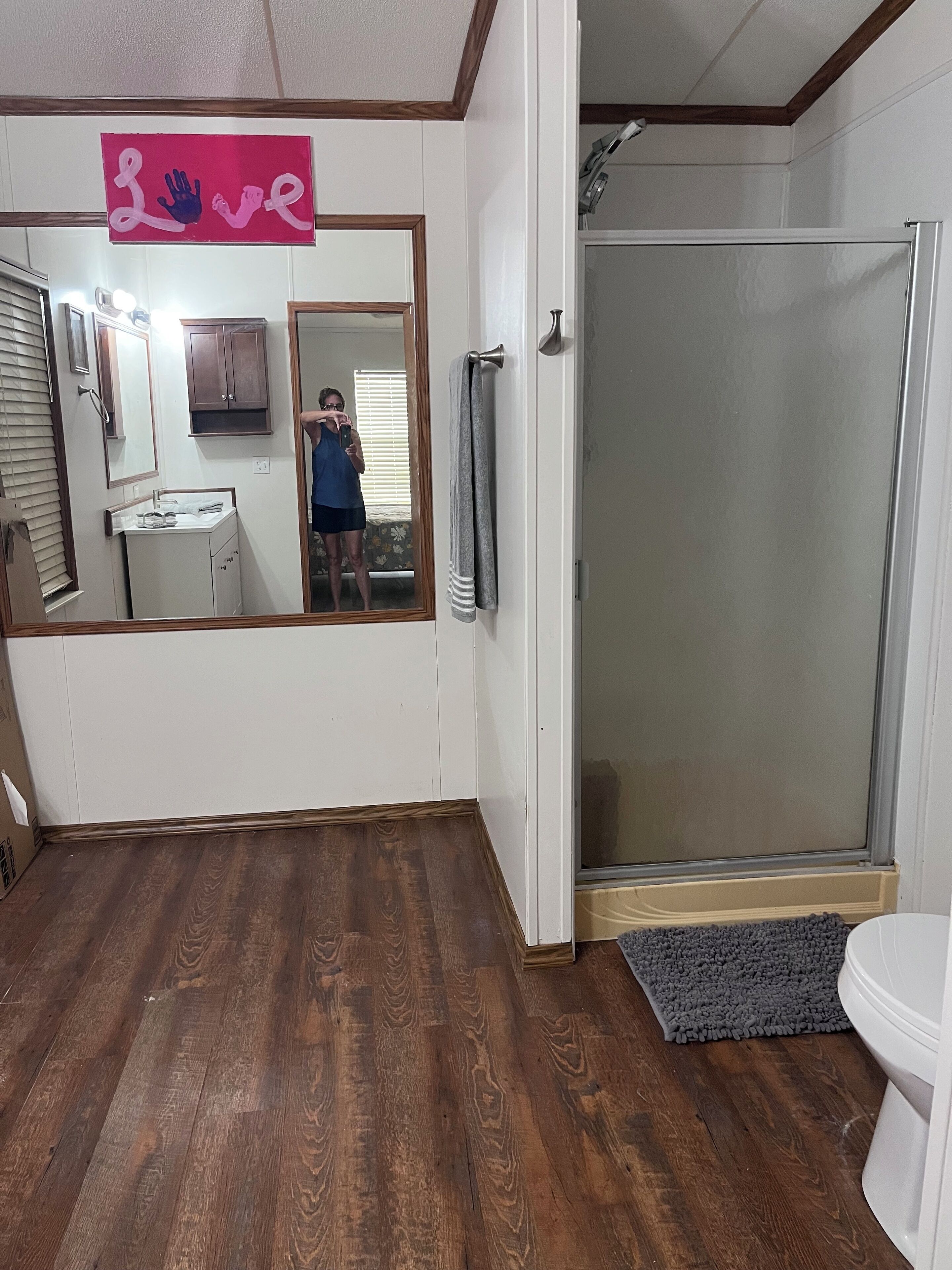 A bathroom with a wooden floor, a shower with a glass door, a sink with a mirror, and a 'Love' sign above the mirror.
