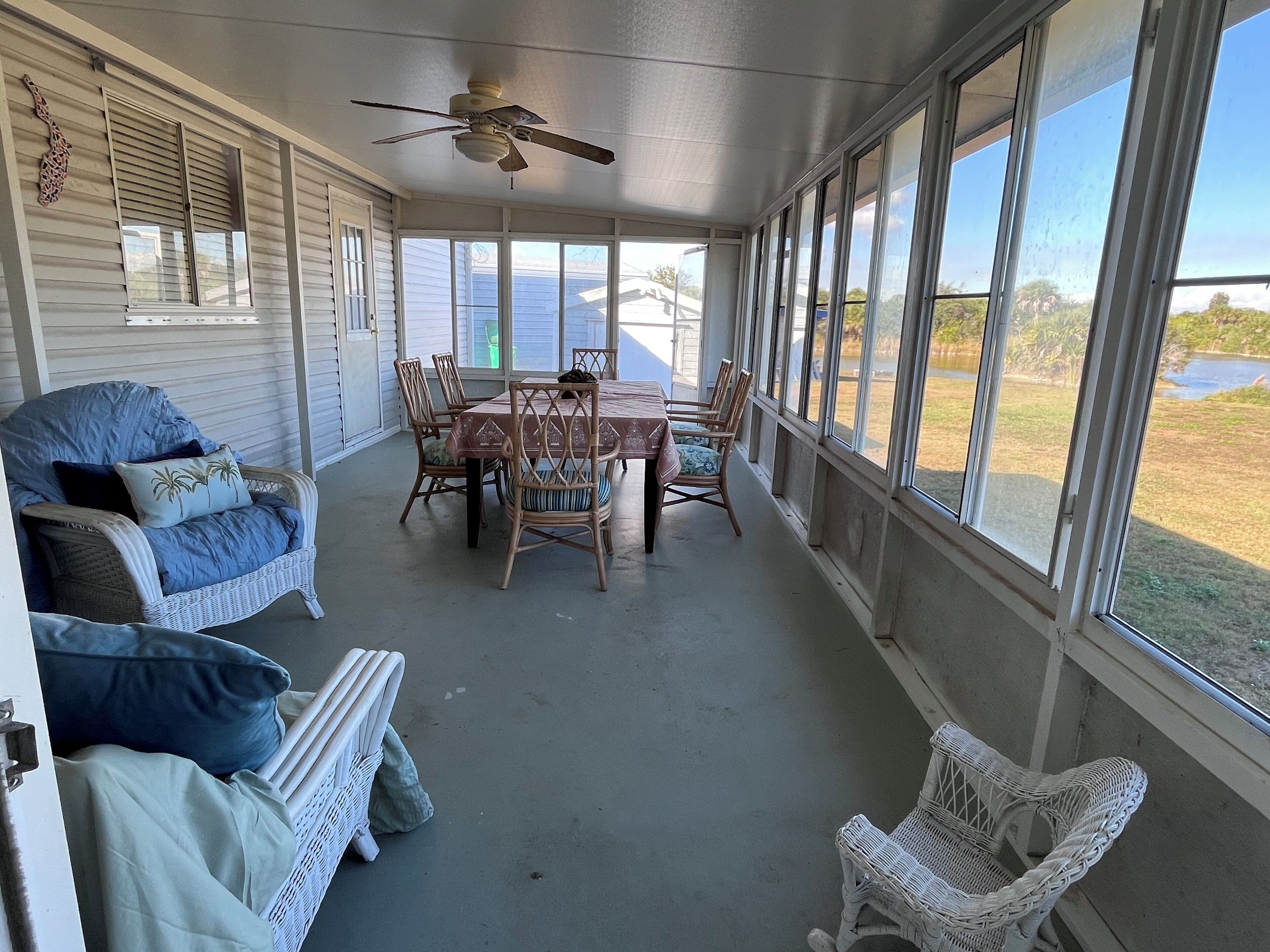 A covered porch with a dining table and chairs, a ceiling fan, and large windows offering a view of a lake and trees.