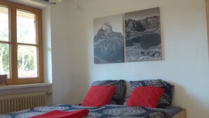 Une chambre à coucher avec un lit en bois, des coussins rouges à motifs, un plaid rouge et deux tableaux représentant des montagnes, accrochés au mur.