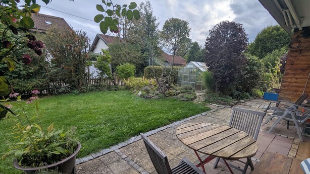 Une terrasse avec une table et des chaises en bois, une pelouse verdoyante et un jardin où poussent différentes plantes, ainsi qu’une serre.