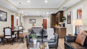 Sala de estar com sofá de couro, mesa de jantar e cadeiras, cozinha com eletrodomésticos e corredor que leva a uma porta.
