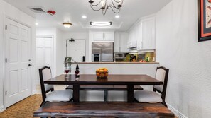 A dining area with a dark wooden table, white upholstered chairs, a chandelier, and a kitchen in the background. - Canyon Lodge at Westgate by Avantstay Park City Mountain Condo With Private Hot Tub + Views + Resort Amenities (Park City)