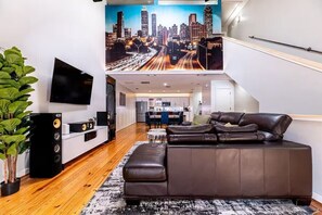 A modern living room with a large brown leather sofa, a flat-screen TV, and a cityscape mural on the wall. - Beltline Townhome / 2-Car Garage Walk to Dining & Groceries (Atlanta)