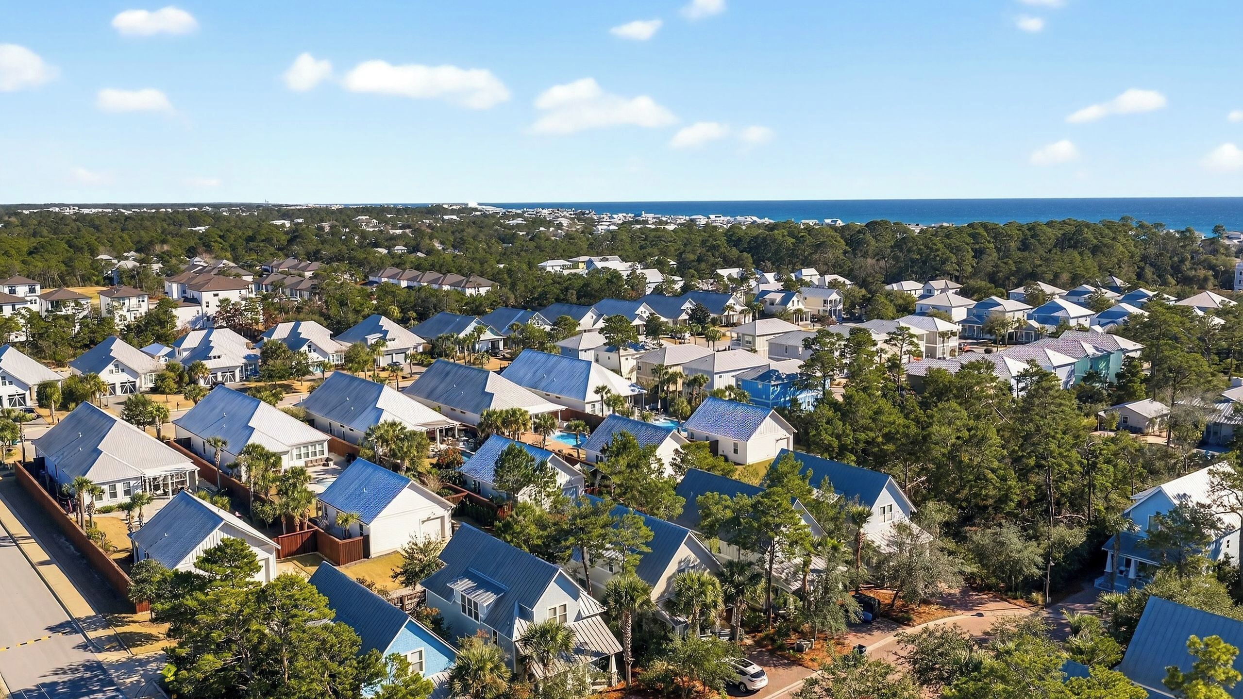 A residential area with houses featuring blue roofs, surrounded by greenery and a clear view of the ocean.