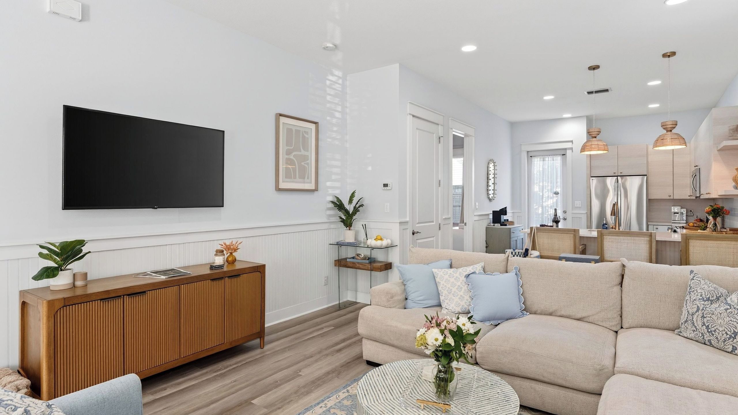 A modern living room with a large flat-screen TV, a wooden console table, a beige sofa with patterned cushions, and a round coffee table.