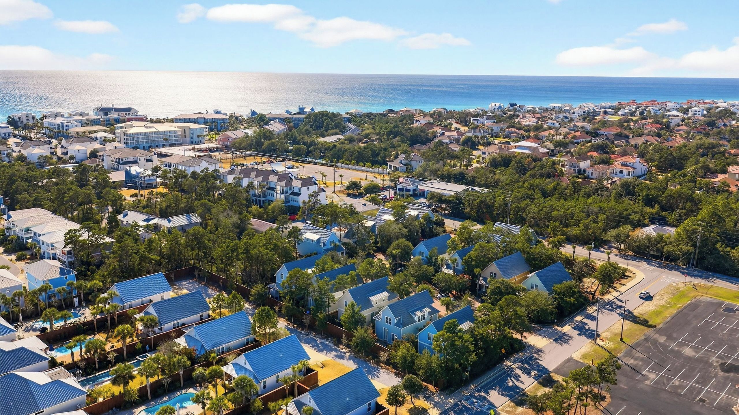 An aerial view of a coastal residential area with houses, a parking lot, and a clear view of the ocean.