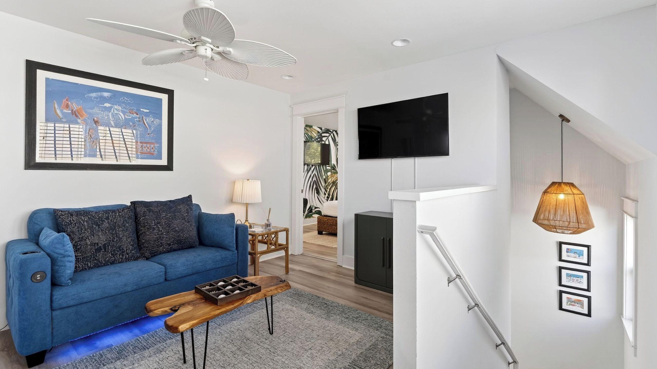 A modern living room with a blue sofa, a wooden coffee table, a ceiling fan, a TV mounted on the wall, and a staircase with a hanging light fixture.