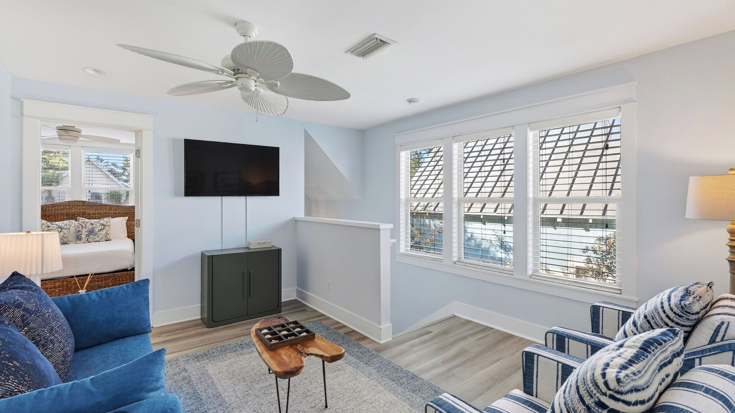 A living room with a ceiling fan, a flat-screen TV, a sofa, a coffee table, and large windows with blinds.