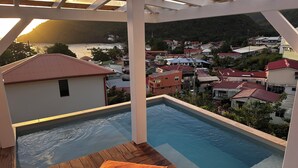 A rooftop pool with a wooden deck and orange cushions, overlooking a coastal town with red-roofed houses and a mountain backdrop.