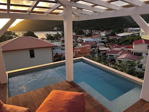 Una piscina en la azotea con cubierta de madera y cojines naranja, con vistas a un pueblo costero de casas de techo rojo y un telón de fondo montañoso.