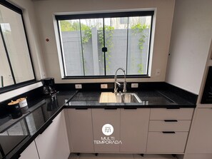 A modern kitchen with a black countertop, a stainless steel sink, and a window with a view of a garden. - Sampaio's House - Gov. Celso Ramos - SC (Governador Celso Ramos)