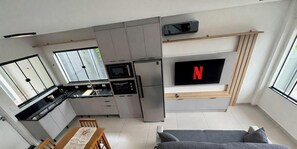 Cocina compacta con horno empotrado, microondas y refrigerador. Un televisor de pantalla plana está montado en la pared, y se aprecia un comedor con mesa y sillas.