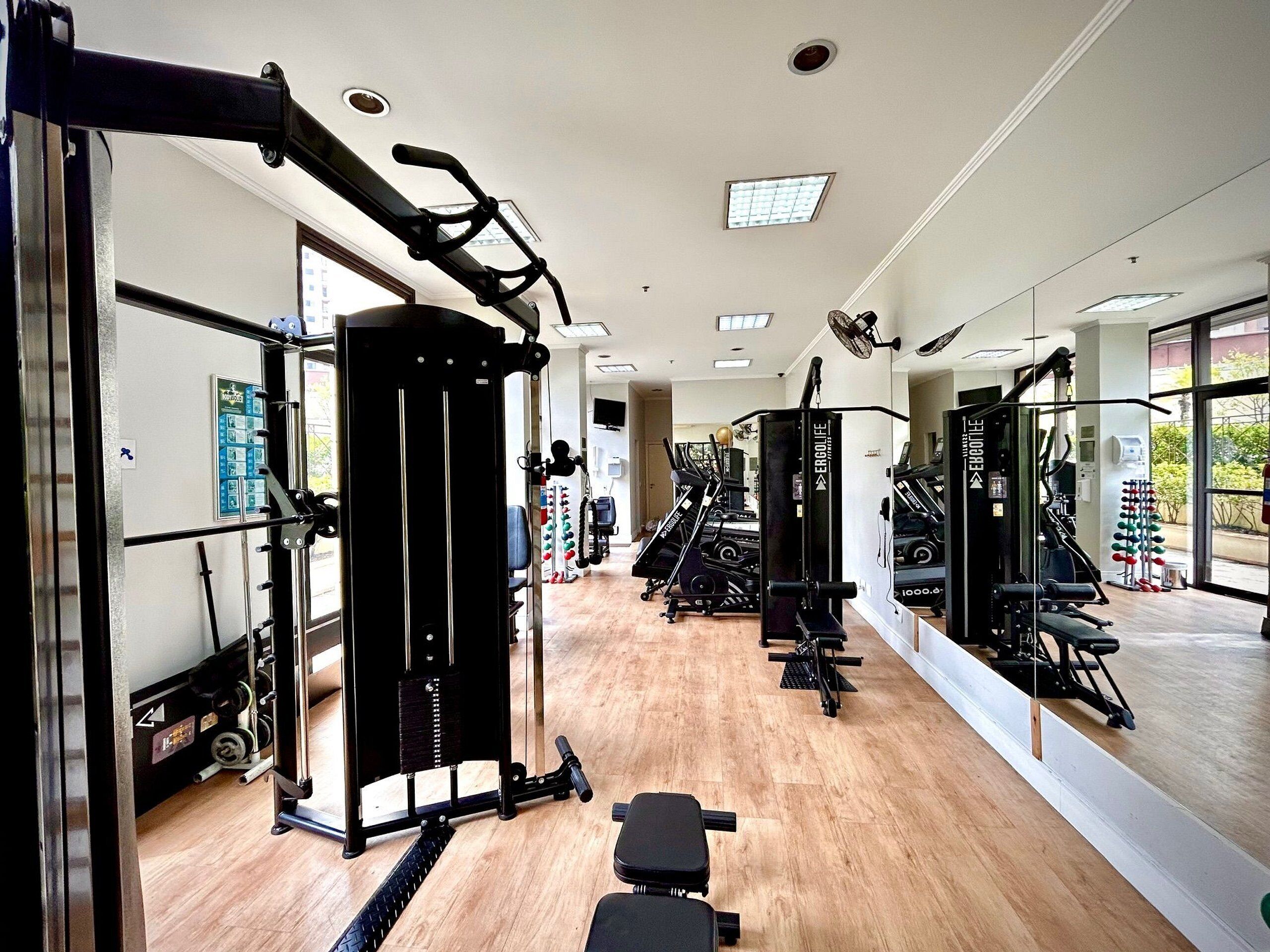 Un gimnasio con diversos equipos de ejercicio, incluyendo máquinas de pesas y equipos de cardio, con amplias ventanas que dejan entrar luz natural.