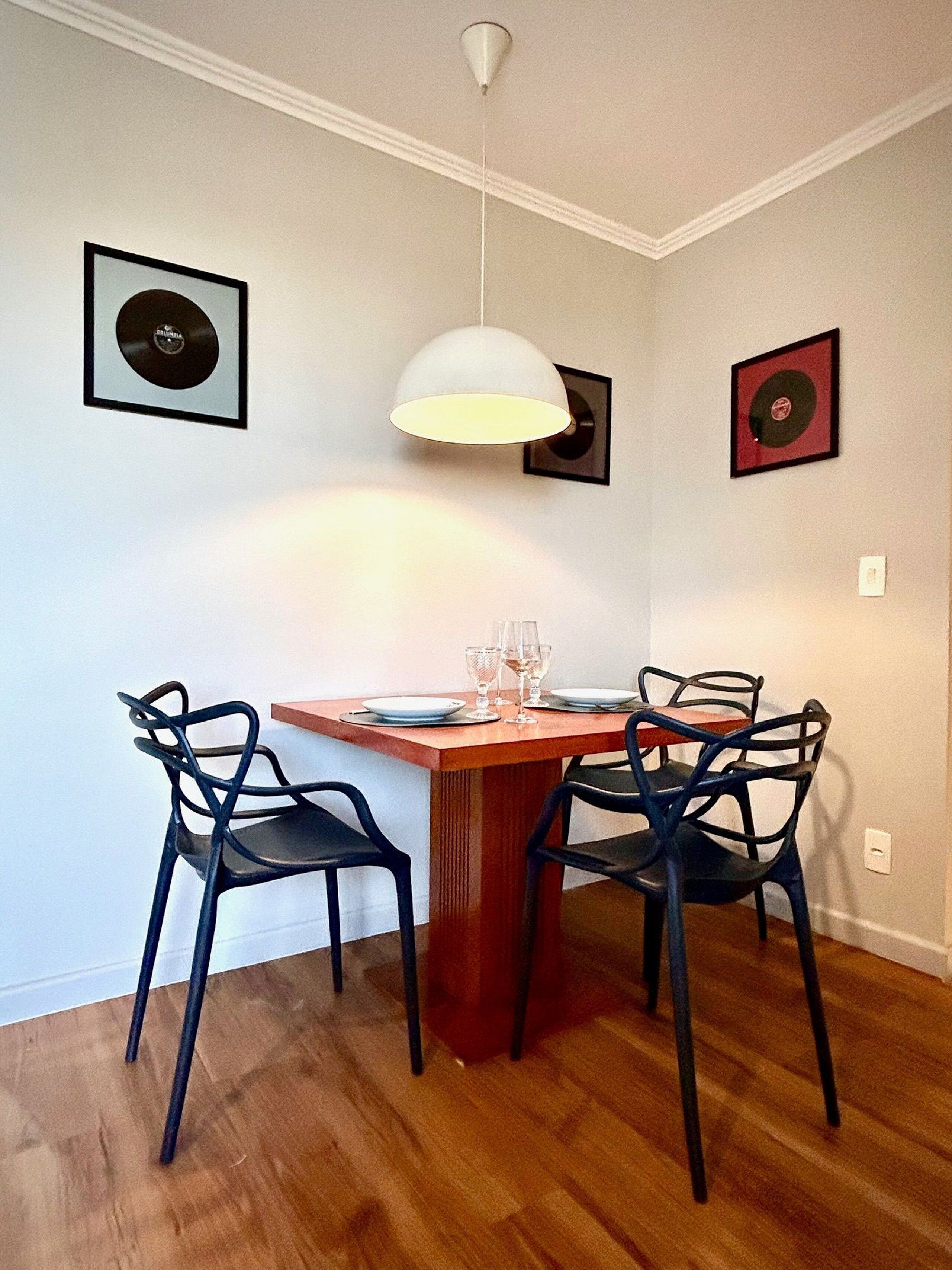 Un coin repas avec une table en bois, quatre chaises noires et un luminaire suspendu blanc. Deux disques vinyle encadrés sont accrochés aux murs.