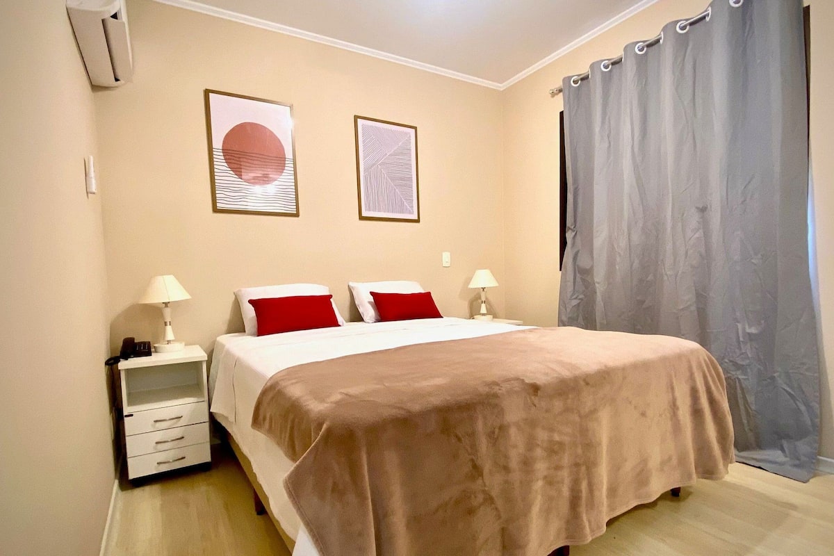 Un dormitorio con cama, dos mesitas de noche, una lámpara, una cortina y dos cuadros en la pared.