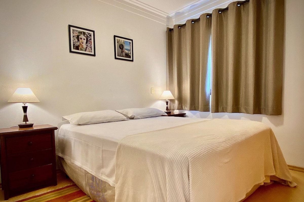 Una cama bien hecha con sábanas blancas, dos lámparas de noche, una alfombra a rayas y dos cuadros en la pared.