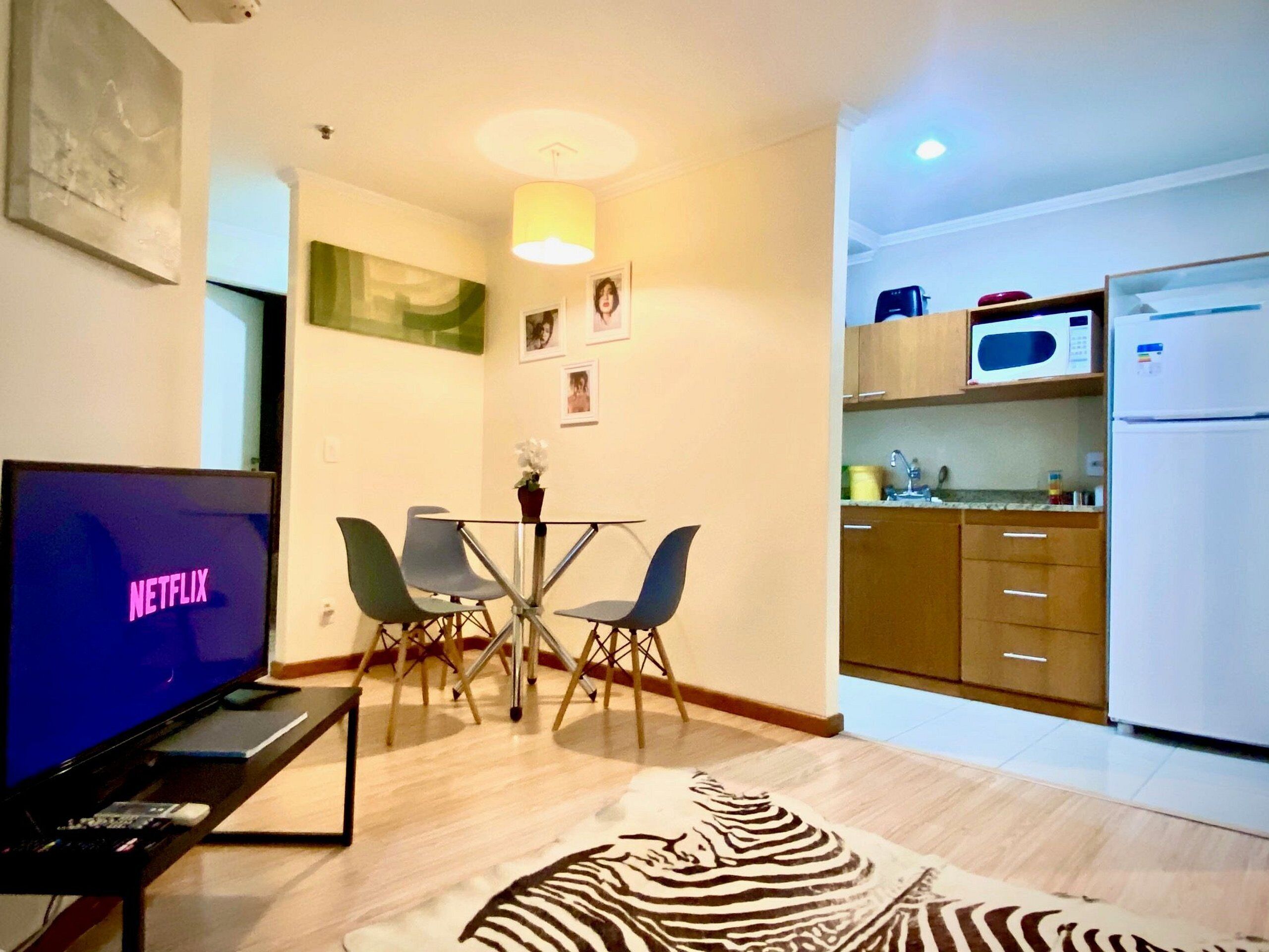 Una sala moderna con un televisor de pantalla plana mostrando 'NETFLIX', una zona de comedor con una mesa de vidrio y sillas, y una cocina con armarios de madera y un refrigerador blanco.