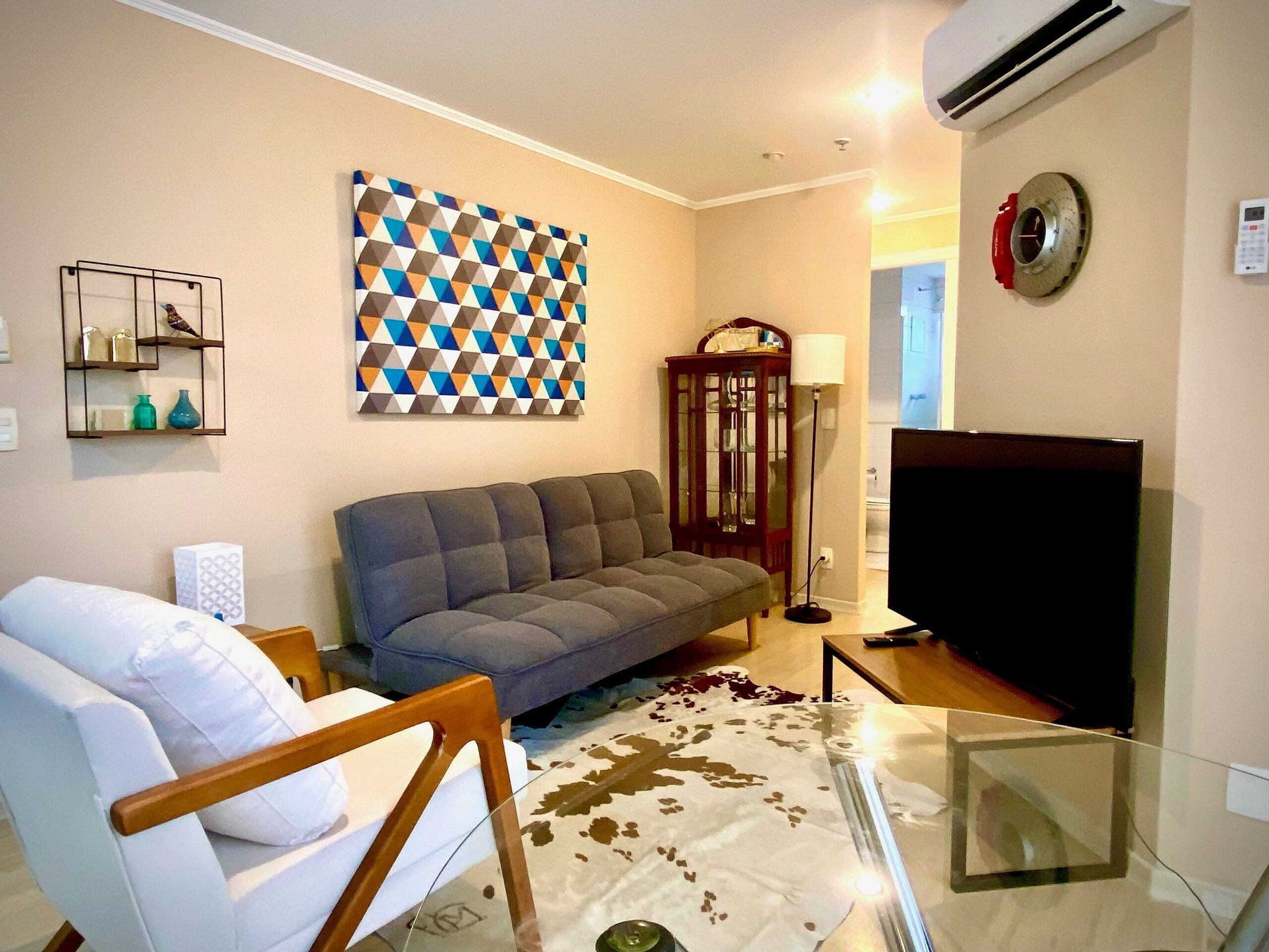 A living room with a sofa, a glass coffee table, a flat-screen TV, a patterned wall hanging, a wooden cabinet, and a floor lamp.