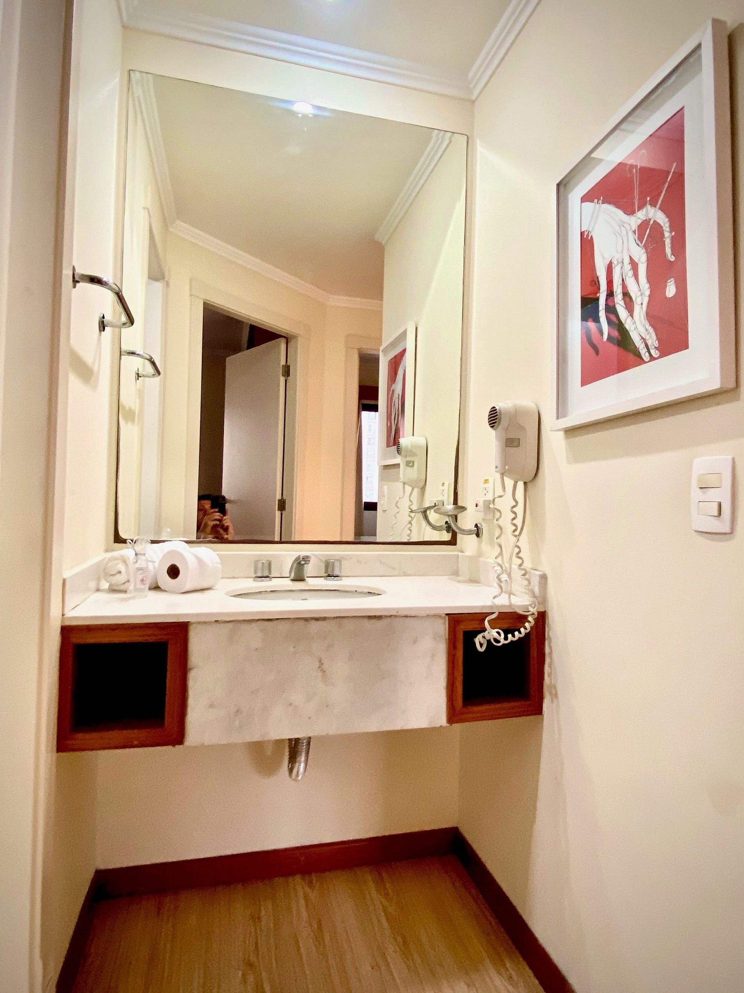 A bathroom with a white sink, a large mirror, a framed artwork on the wall, and a towel dispenser.