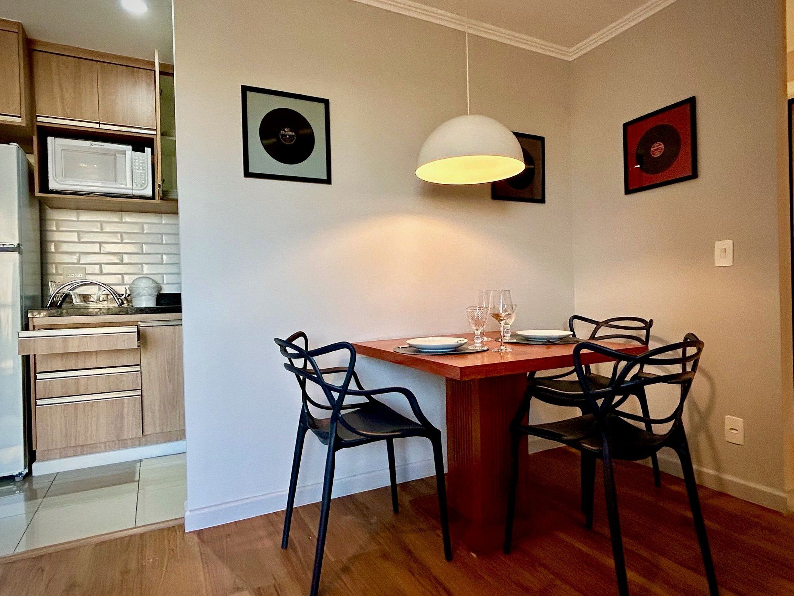 Cozinha compacta com micro-ondas, armários de madeira e área de refeições com mesa e cadeiras. Há discos de vinil emoldurados na parede.