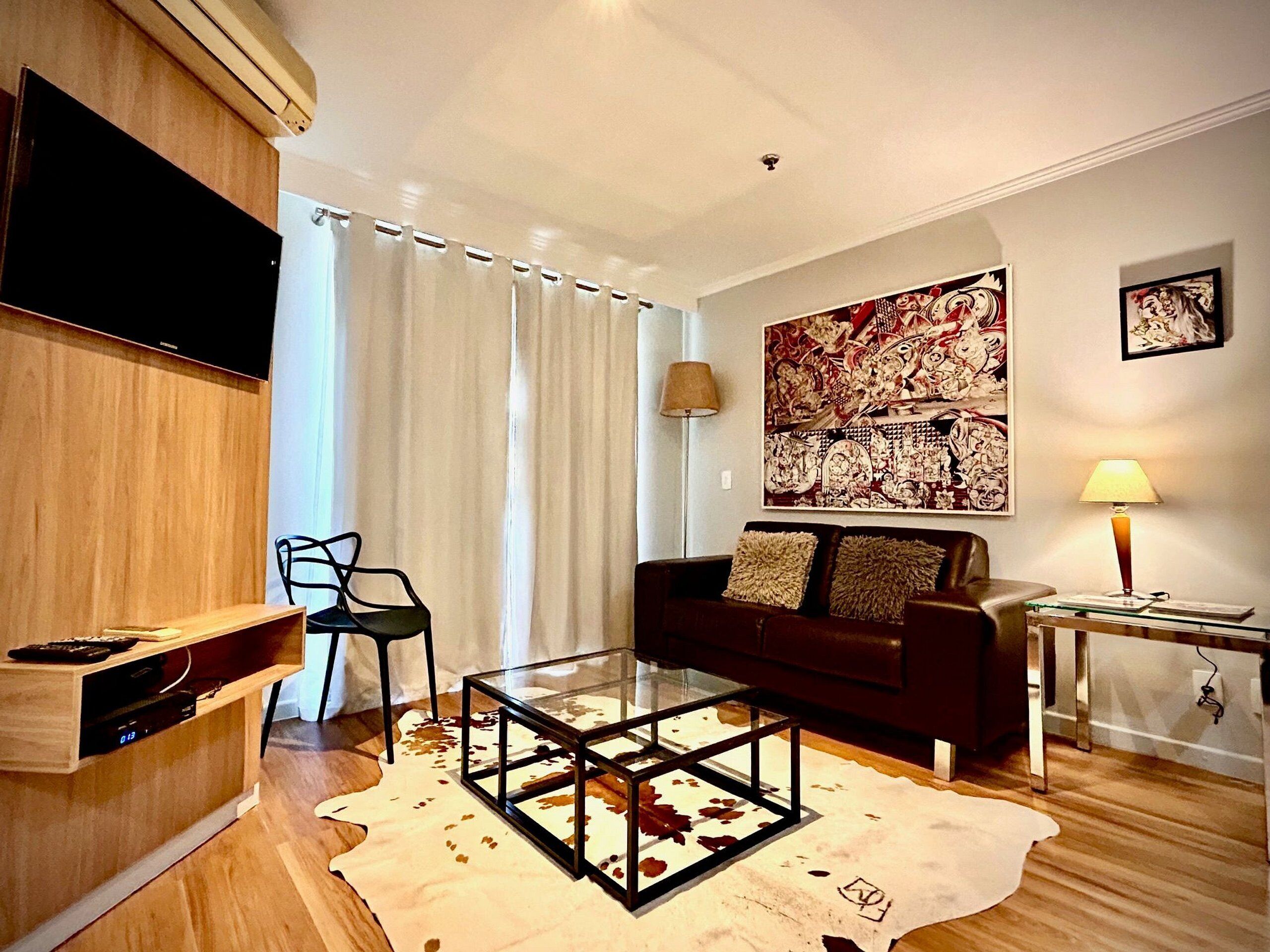 Ein modernes Wohnzimmer mit einem Glastisch, einem dunkelbraunen Sofa, einem an der Wand montierten Flachbildfernseher und einem großen abstrakten Gemälde.