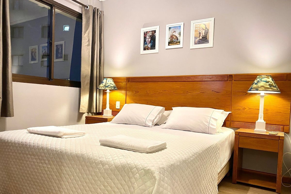 Una cama bien hecha con sábanas y almohadillas blancas, dos lámparas de noche y tres cuadros en la pared.