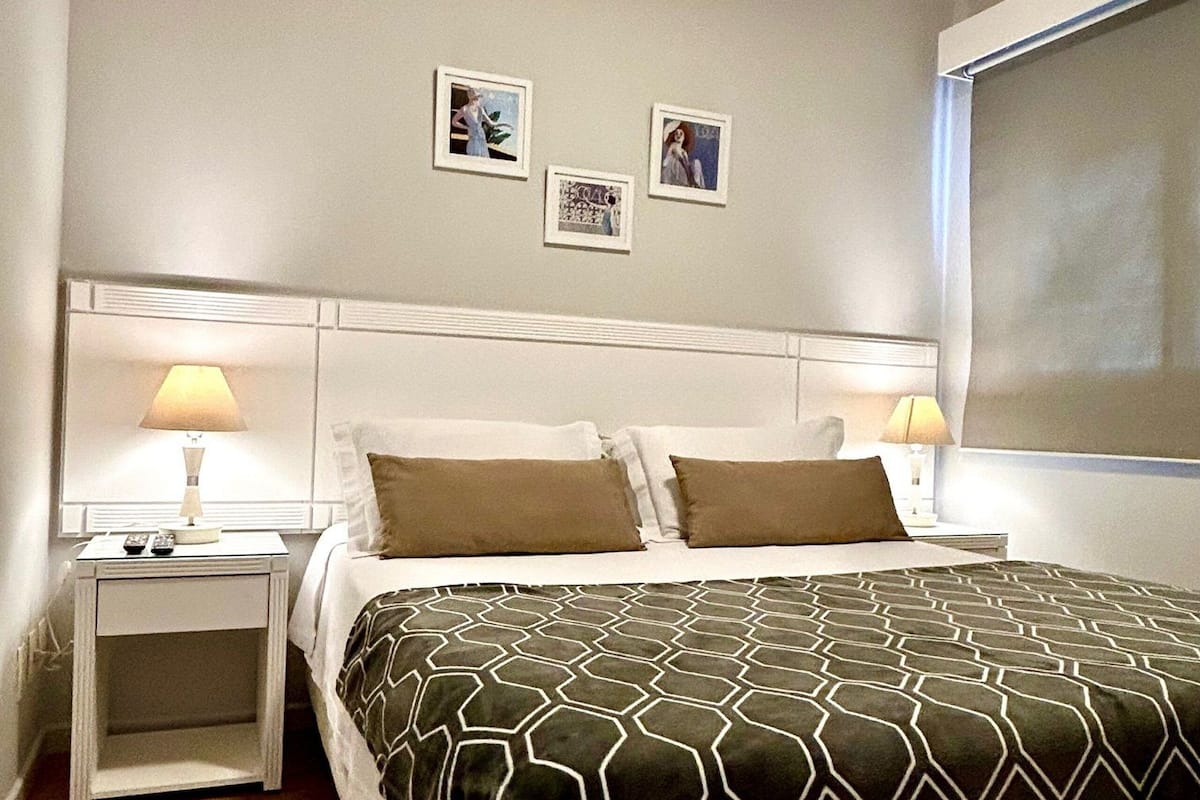 Habitación de hotel ordenada con una cama grande, dos mesitas de noche con lámparas, un cabecero y tres cuadros enmarcados en la pared.
