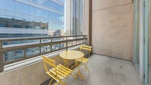 Un balcon avec une table ronde et des chaises jaunes, un mur en briques et, au fond, un grand immeuble en verre.