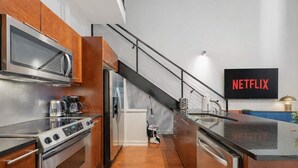 Een moderne keuken met roestvrijstalen apparatuur, houten kasten, een ingebouwde oven en een groot eiland met een spoelbak. Een trap leidt naar een hoger gelegen verdieping, en een aan de muur bevestigde televisie toont het woord 'NETFLIX'.
