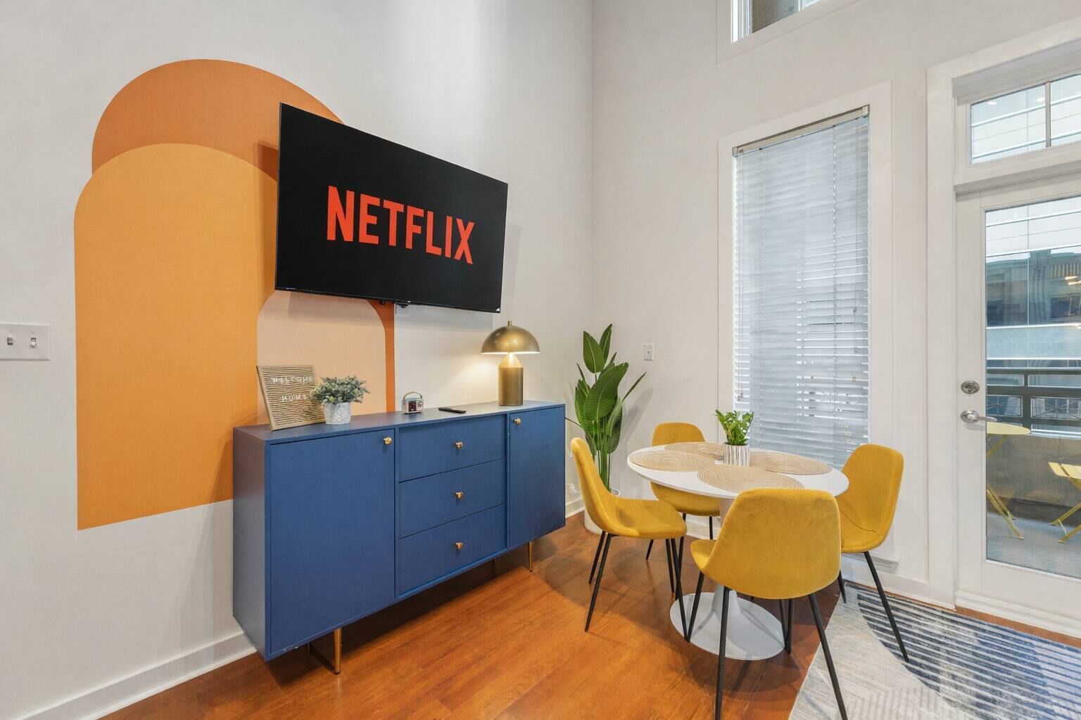 A modern living room with a blue cabinet, a round dining table with yellow chairs, a TV mounted on the wall, and a large window with blinds.
