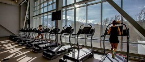 Une salle de sport avec des tapis de course disposés en rangées, de grandes fenêtres et une vue sur les montagnes.