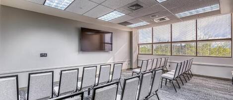 A conference room with rows of chairs, a flat-screen TV mounted on the wall, and large windows with blinds.