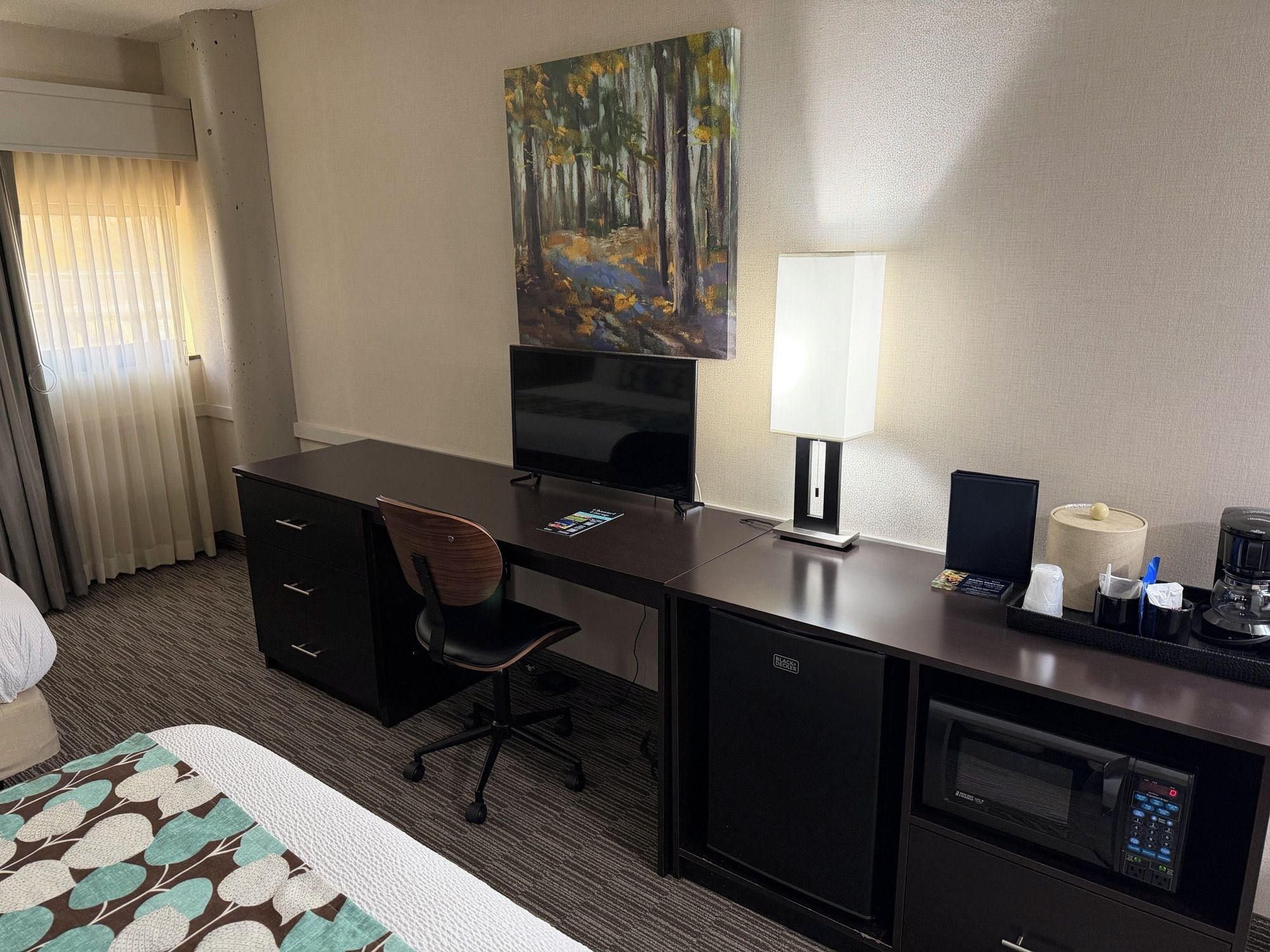A hotel room with a desk, chair, television, lamp, coffee maker, and a painting on the wall.