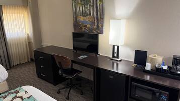 A hotel room with a desk, chair, television, lamp, coffee maker, and a painting on the wall.