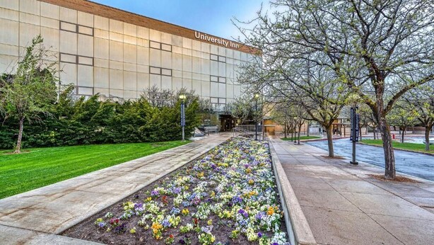 Un bâtiment de l’hôtel universitaire doté d’une grande façade en verre, d’un jardin aménagé avec des fleurs aux couleurs vives et d’un chemin bordé d’arbres.