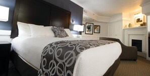 A hotel room with a large bed, a nightstand, a chair, a fireplace, and framed artwork on the wall. - University Inn (Logan)