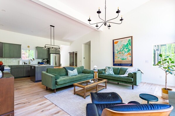 A modern living room with green sofas, a coffee table, and a dining area with a kitchen in the background.