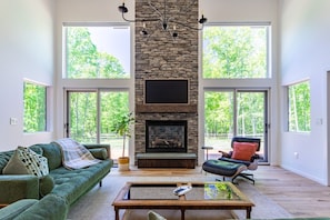 A spacious living room with a stone fireplace, large windows, a green sofa, and a modern chair.