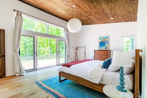 A bedroom with a wooden ceiling, a large window with curtains, a bed with white bedding, a blue rug, and a bedside table with a lamp.